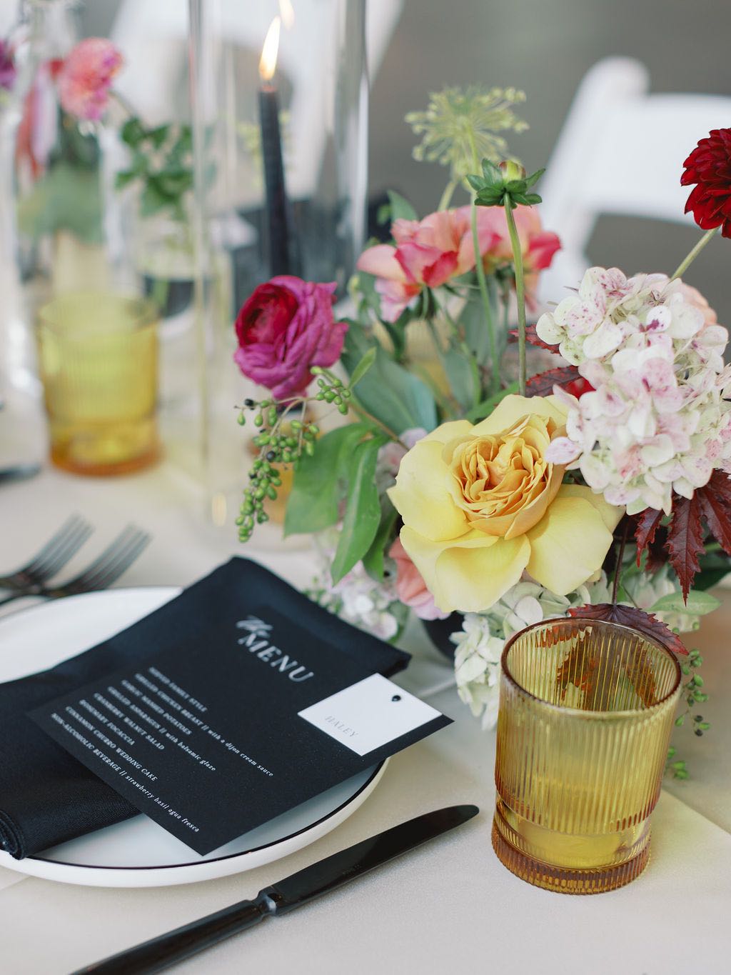 Wedding place setting with black napkin, menu and flatware and amber glassware. Colorful flowers down the center