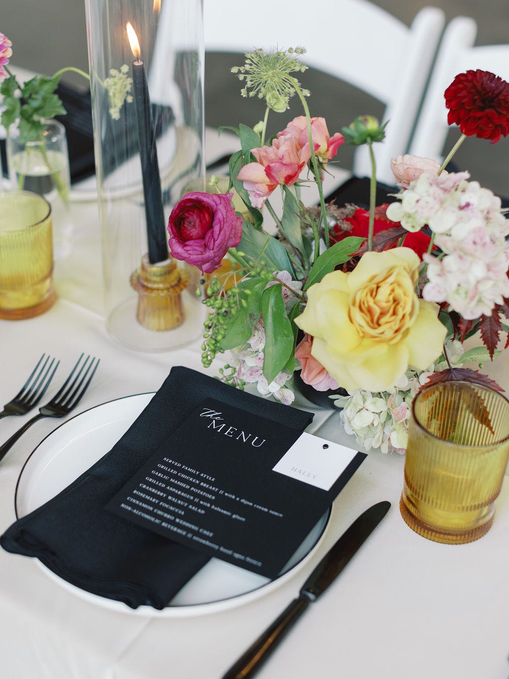Wedding place setting with black napkin, menu and flatware and amber glassware. Colorful flowers down the center