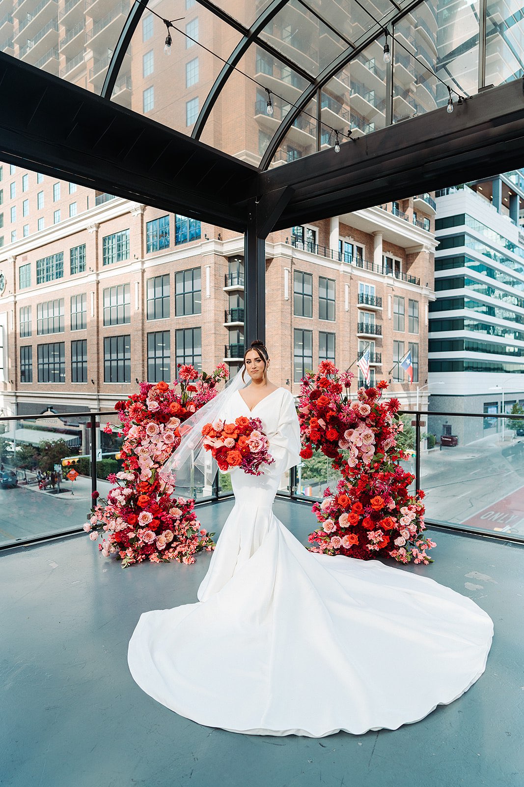 Bride holding red and pink bouquet infront of Asymmetrical wedding floral arch with mostly red and pink flowers, overlooking downtown Austin