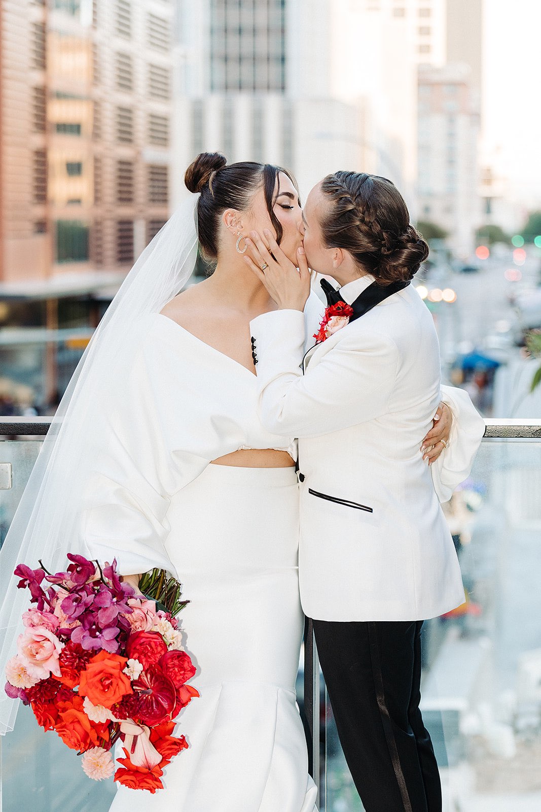 Brides kissing on rooftop wedding venue while holding red bouquet