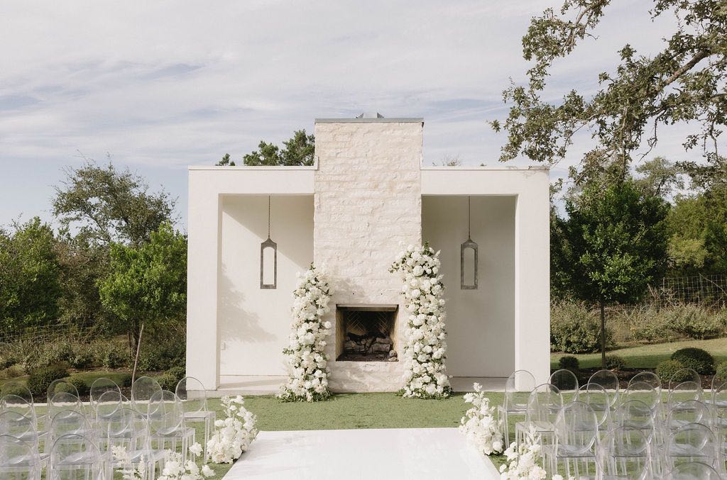 Outdoor wedding ceremony with white floral arches, a white acrylic aisle runner and clear ghost chairs 