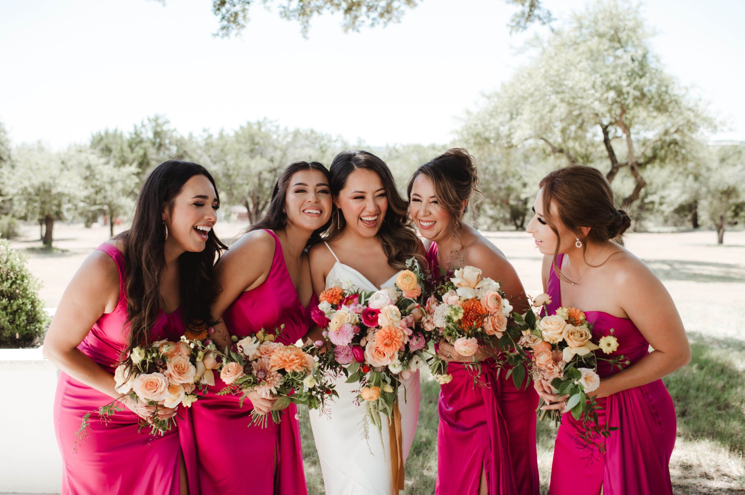 Bride laughing with her bridesmaids who are wearing pink dresses and holding summery peach bouquets