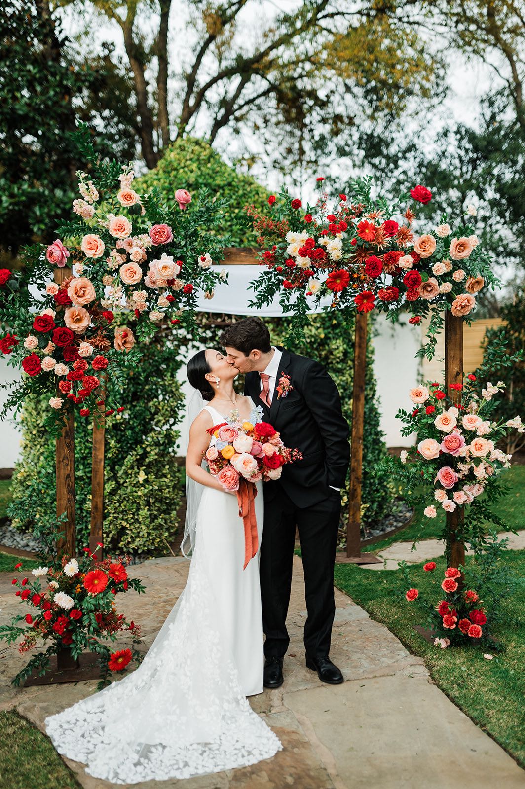Newlyweds kiss under a chuppah covered in colorful flowers in shades of blush, peach and red. 